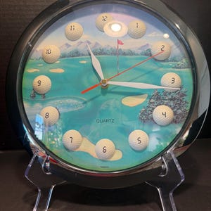 May include: A round wall clock with a black frame and a golf course scene on the face. Twelve white golf balls serve as hour markers, each labeled with a black number. The clock features white hour and minute hands and a thin red second hand.