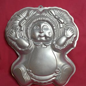May include: A vintage silver-colored metal cake pan in the shape of a doll. The pan features a detailed design of a doll's face, hair, and clothing. It has a loop for hanging and is likely used for baking decorative cakes.