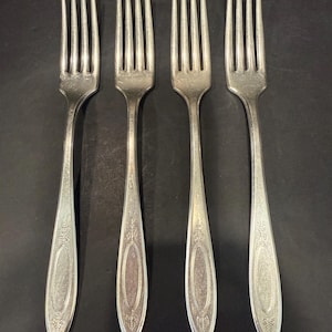 May include: A set of four silver-toned metal forks arranged in a row. Each fork features a four-tined head and a handle decorated with an embossed oval center panel surrounded by intricate leaf-like patterns at the top and bottom ends.