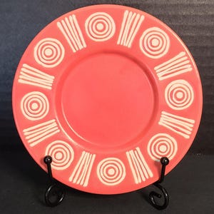 May include: A round, coral-colored plate featuring a repeating pattern of white concentric circles and sets of three vertical lines around the rim. The plate is displayed on a black metal stand, showcasing a smooth, matte-textured surface.