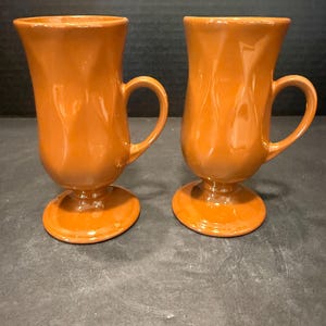 May include: Two matching orange ceramic mugs with a glossy finish. Each mug features a tall, flared body with a subtle vertical rippled texture, a curved side handle, and a circular pedestal base that elevates the cup from the surface.