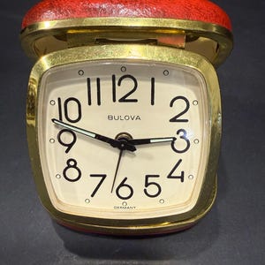 May include: A vintage Bulova travel alarm clock with a red textured leather-like case and a gold-tone metal frame. The square clock face is off-white with black Arabic numerals, black hands with luminous accents, and the word Germany at the bottom.