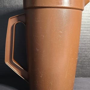 May include: A brown plastic pitcher featuring a textured surface, a sturdy side handle, and a molded pouring spout. The cylindrical body has a slightly tapered shape with a series of vertical notches along the base, showing signs of surface wear.