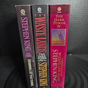 May include: Three paperback books from The Dark Tower series by Stephen King, published by Plume. The spines show titles: The Drawing of the Three, The Waste Lands, and Wizard and Glass. The covers feature black, dark purple, and magenta colors.
