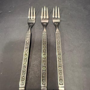 May include: Three silver-toned metal forks arranged vertically. Each fork features a long, flat handle decorated with a repeating embossed pattern of small circles, hearts, and floral shapes. The tines are slender and pointed, with a polished finish.