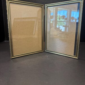 May include: A double-hinged picture frame featuring two rectangular vertical openings with thin, metallic gold-toned borders. The frames are connected by a metal hinge, allowing them to stand upright in an open, angled position with glass panes.