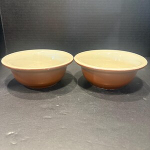 May include: Two round ceramic bowls featuring a smooth, glossy cream-colored interior and a contrasting matte terracotta-orange exterior. Each bowl has a wide, slightly flared rim and a rounded base, presented side by side in a simple arrangement.