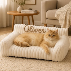 May include: A cream-colored, ribbed pet sofa featuring a personalized name tag that reads Oliver in cursive script. A long-haired ginger cat is resting on the soft, textured surface of the bed, which has a backrest and two side armrests.