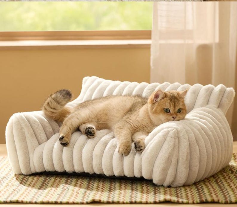 May include: A cream-colored, plush pet sofa with a ribbed, tufted texture and rounded armrests. A light brown tabby cat with green eyes is lying across the center of the soft, cushioned seat, resting its body against the high, padded backrest.