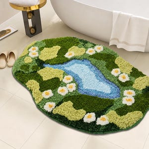 May include: An oval-shaped tufted rug featuring a landscape design with a central light blue pond, surrounded by various shades of green grass and small white flowers with yellow centers. The rug has a textured, plush pile surface throughout.