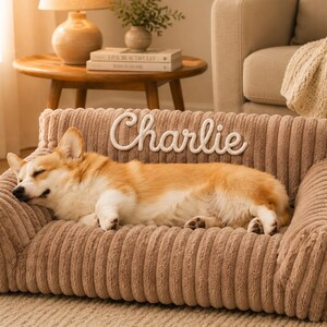 May include: A tan, corduroy-textured pet sofa featuring a high backrest with the name Charlie embroidered in white script lettering. The sofa has a rectangular base with cushioned armrests and a backrest, providing a soft surface for a dog.