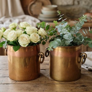 May include: Two antique copper vases with brass handles. One vase holds white roses, the other holds eucalyptus branches. The vases sit on a rustic wooden table, creating a vintage aesthetic.