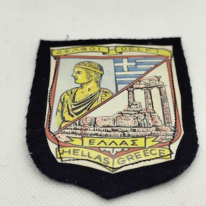May include: A navy blue patch with a shield design featuring a yellow bust, Greek flag, and ancient ruins. The patch includes the text "DELPHI", "ΕΛΛΑΣ", and "GREECE". The patch has a black border.