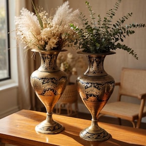 May include: Two decorative gold vases with black patterns, each holding dried flowers and greenery. The vases are displayed on a wooden table. The vases are approximately 30 cm tall, creating an elegant display.