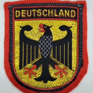 May include: Embroidered patch featuring the word "DEUTSCHLAND" in gold lettering. The patch has a black eagle emblem with red accents on a yellow background, framed by a black border and a red felt edge.