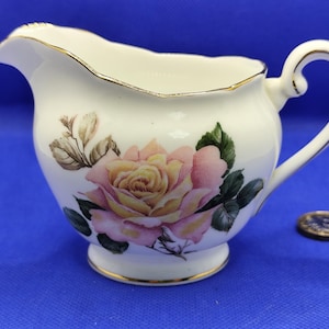 Può includere: Una crema in porcellana bianca con un disegno di rosa gialla e rosa. La crema ha un manico curvo e un bordo dorato attorno al bordo e alla base. Una moneta è accanto alla crema.