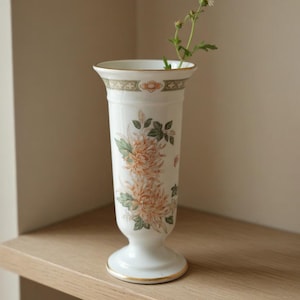 Puede incluir: Un jarrón de flores vintage blanco con borde dorado y diseño floral. El jarrón presenta crisantemos naranjas y verdes. El jarrón está hecho en Inglaterra y contiene una pequeña ramita de flores.