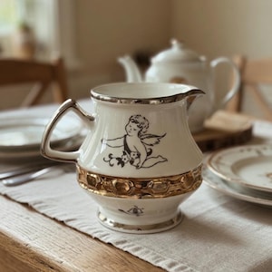 Könnte beinhalten: Cremefarbener Keramik-Sahnekännchen mit goldenem Rand und Griff. Das Kännchen zeigt eine schwarze Illustration eines Engels und goldene Details am Boden. Eine weiße Teekanne ist im Hintergrund zu sehen, was auf ein Teeservice hindeutet.