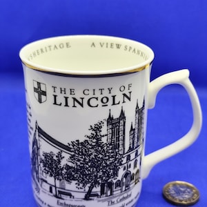 May include: White ceramic mug featuring a black and white illustration of a cathedral and the text "THE CITY OF LINCOLN". The mug has a gold rim and a white handle. A coin is next to the mug.