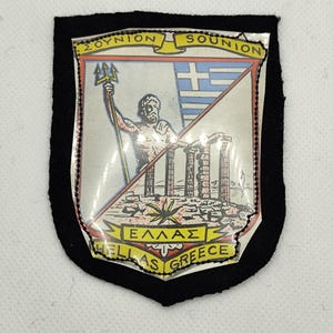May include: A shield-shaped patch with a black border. The center features a colourful illustration of a Greek scene, including a figure with a trident, the Greek flag, and the text "Sounion" and "Greece".