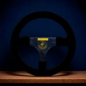 May include: A black suede steering wheel featuring a carbon fibre centre spoke with the yellow MOMO logo at the top and a yellow Lotus Team Lotus emblem in the centre. The steering wheel has a circular rim and a three-spoke design.
