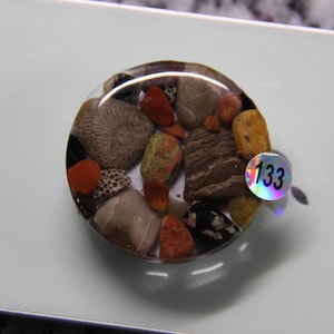 May include: A circular resin paperweight containing a collection of small, multi-colored natural stones in shades of red, brown, gray, and yellow. A small, round, holographic sticker with the black number 133 is attached to the right side.