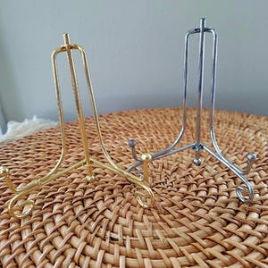 May include: Two metal display stands. One is gold-colored, the other is silver. Both have a three-legged base and a curved top to hold a plate or other item. The stands are on a woven, brown surface.