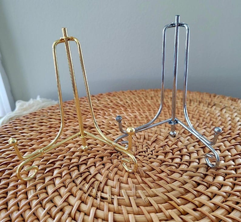 May include: Two metal display stands. One is gold-colored, the other is silver. Both have a three-legged base and a curved top to hold a plate or other item. The stands are on a woven, brown surface.