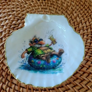 May include: A white scallop shell dish with a gold-painted rim featuring a colorful illustration of a cartoon character with a white beard and sunglasses wearing a green shirt, floating in a blue floral inner tube while holding a glass of beer.