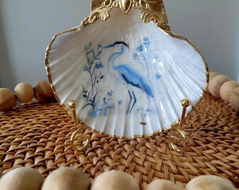 May include: A decorative seashell-shaped dish with a gold-painted rim and stand. The dish features a hand-painted blue heron and floral design. The dish is displayed on a woven surface with wooden beads.