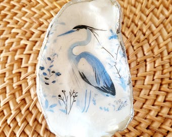 May include: A white oyster shell featuring a hand-painted blue and black heron illustration. The bird is depicted in profile surrounded by delicate blue floral accents. The edges of the shell are finished with a metallic silver trim, creating a decorative piece.