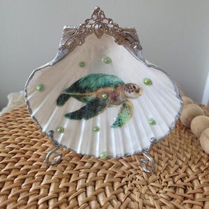 May include: A decorative seashell trinket dish featuring a painted sea turtle. The white shell is trimmed with silver and adorned with green beads. The dish is supported by a silver metal stand, creating a coastal theme.