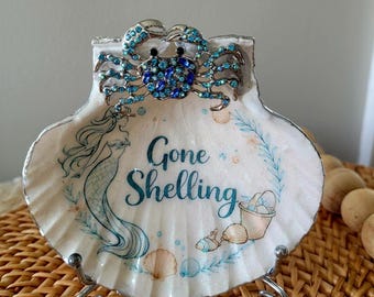 May include: A decorative seashell-shaped trinket dish with a crab embellishment. The dish features a mermaid illustration and the words "Gone Shelling" in teal. The crab is adorned with blue and teal rhinestones. The dish is supported by a silver metal stand.