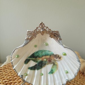 May include: A decorative seashell trinket dish featuring a painted sea turtle. The white shell has a silver trim and is adorned with green beads. The dish is supported by a silver metal stand and has a decorative silver crown.