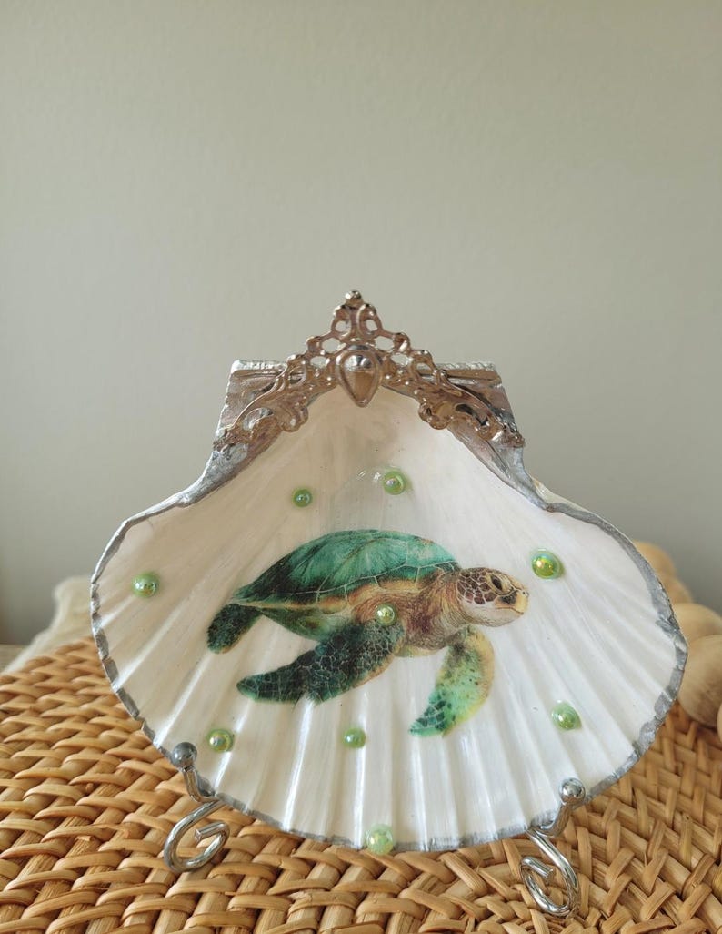 May include: A decorative seashell trinket dish featuring a painted sea turtle. The white shell has a silver trim and is adorned with green beads. The dish is supported by a silver metal stand and has a decorative silver crown.