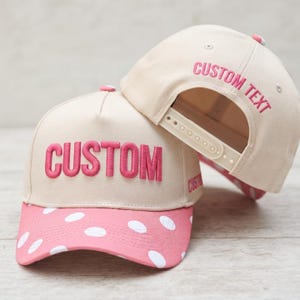 May include: Two beige baseball caps with pink polka-dot brims. The front cap features the word CUSTOM in pink embroidery. The back cap shows the text CUSTOM TEXT embroidered in pink above the adjustable plastic snapback closure strap.