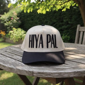 May include: A beige and navy blue baseball cap with the text "HIYA PAL" embroidered in black. The cap is sitting on a wooden table outdoors, with greenery in the background.