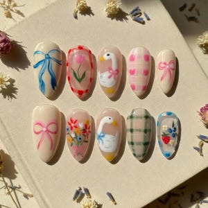 May include: A set of ten almond-shaped false nails with various hand-painted designs. The designs include bows, flowers, a goose, and plaid patterns in pastel colors like pink, blue, and green. The nails are arranged on a light-colored surface.