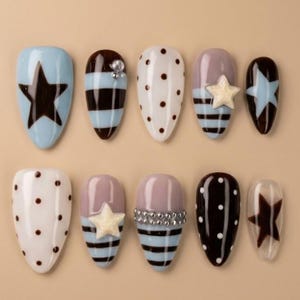May include: A set of ten artificial nails in various designs. The nails feature a light blue base with a dark star, brown and blue stripes, white with brown polka dots, and a mauve base with a white star.