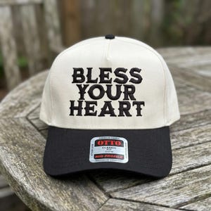 May include: A cream-colored baseball cap with a black brim. The phrase "BLESS YOUR HEART" is embroidered in bold black letters across the front. The cap has a mid-profile design and a tag that reads "OTTO CLASSIC OSFM".