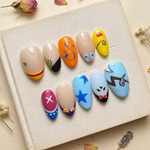 May include: A set of ten oval-shaped, painted artificial nails. Each nail features a unique design, including a cross, a star, and various cartoon-inspired illustrations. Colors include blue, orange, yellow, and nude tones. The nails are arranged on a white surface.