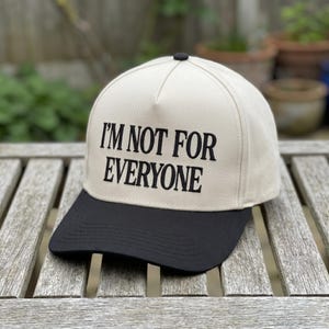 May include: A beige and black baseball cap with the text "I'M NOT FOR EVERYONE" in black. The cap has a black brim and a black button on top. The hat is sitting on a wooden surface.