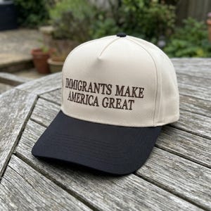 May include: A beige and black baseball cap with the embroidered text "IMMIGRANTS MAKE AMERICA GREAT". The cap has a black brim and a black button on top. The hat is sitting on a wooden surface.