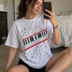 Può includere: T-shirt bianco con un disegno di tastiera di pianoforte, una striscia rossa e note musicali. La grafica della tastiera in bianco e nero è centrata, con note musicali sparse. Un capo d'abbigliamento casual a tema musicale.