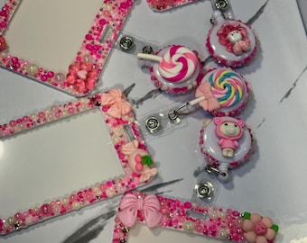 May include: A collection of rectangular badge holders and circular retractable badge reels decorated with pink beads, rhinestones, and charms including bows, flowers, cherries, lollipops, and a small bear figure in a pink hooded outfit.