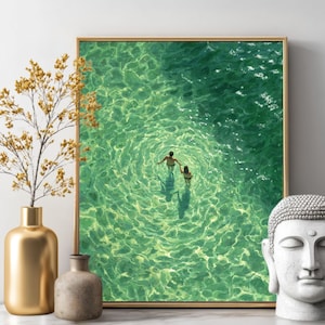 May include: Framed art print featuring two figures in turquoise water, holding hands. The artwork is in a gold frame and is displayed with a gold vase, a small stone vase, and a white Buddha head.