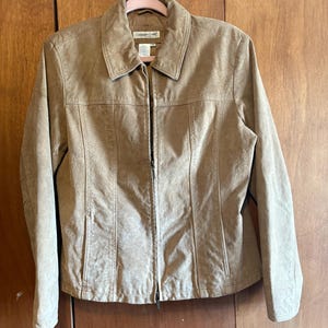 May include: A tan suede jacket with a collared neckline and a full-length zipper. The jacket has vertical seams on the front and is hanging on a pink hanger. The jacket is a neutral color and appears to be made of a soft material.