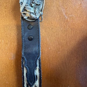May include: A black leather belt with a decorative silver-colored buckle featuring a detailed relief design. The belt has a woven pattern in cream and black, and is secured with two black snaps.