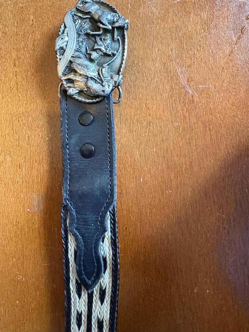 May include: A black leather belt with a decorative silver-colored buckle featuring a detailed relief design. The belt has a woven pattern in cream and black, and is secured with two black snaps.