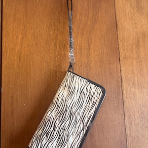 Puede incluir: Una cartera rectangular con marco negro y un estampado de rayas de cebra en blanco y negro. La cartera cuelga de una correa negra sujeta a un gancho metálico.